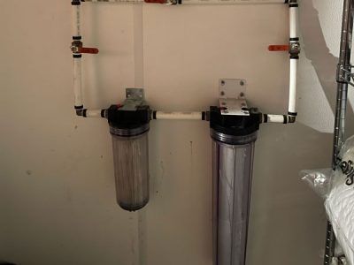 Water filtration system with two clear canisters connected by white PVC piping and valves, mounted on a white wall.