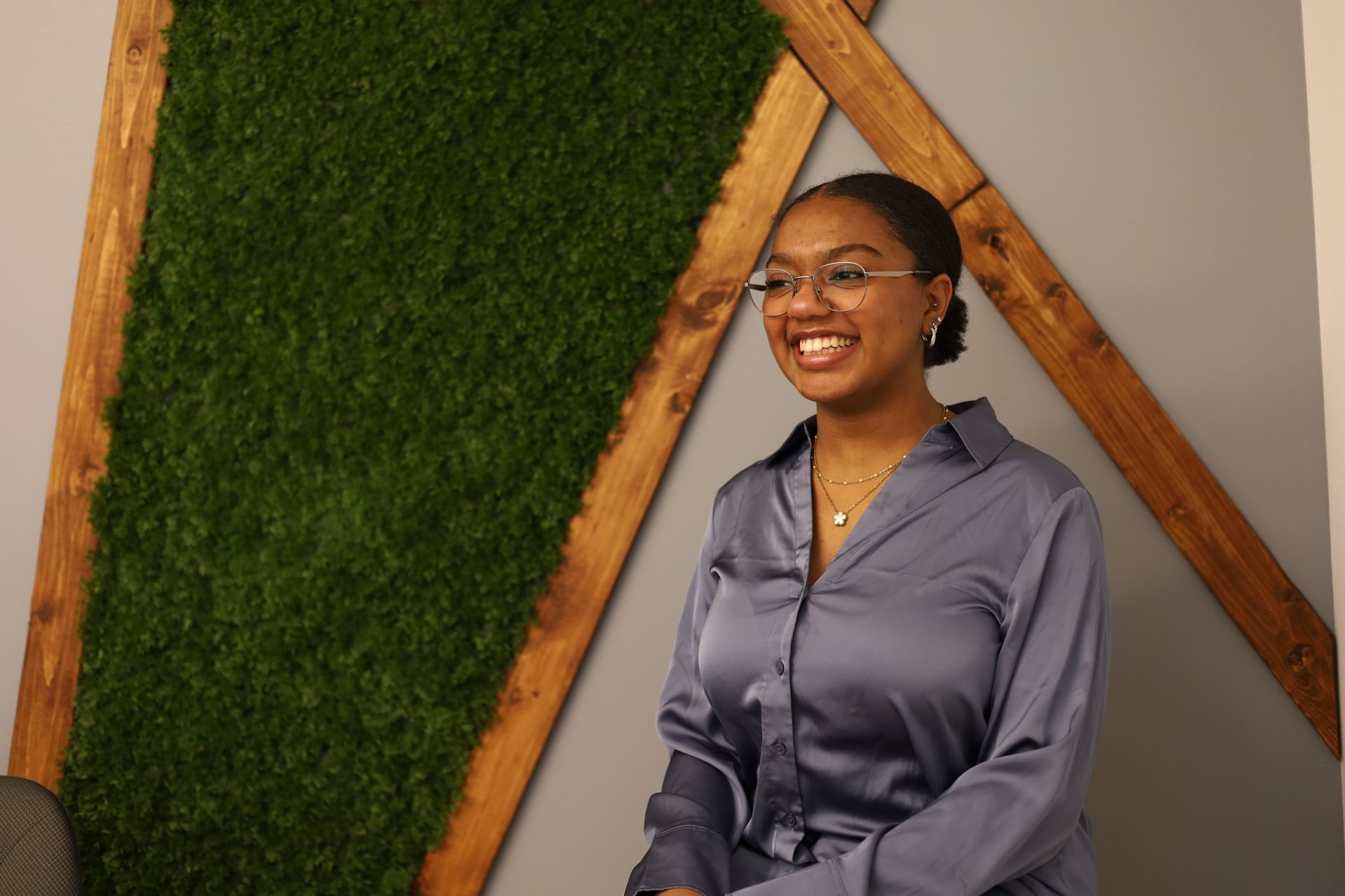 Femme à lunettes, bras croisés, souriante. Elle porte un blazer, une chemise blanche et un jean devant un fond bleu.