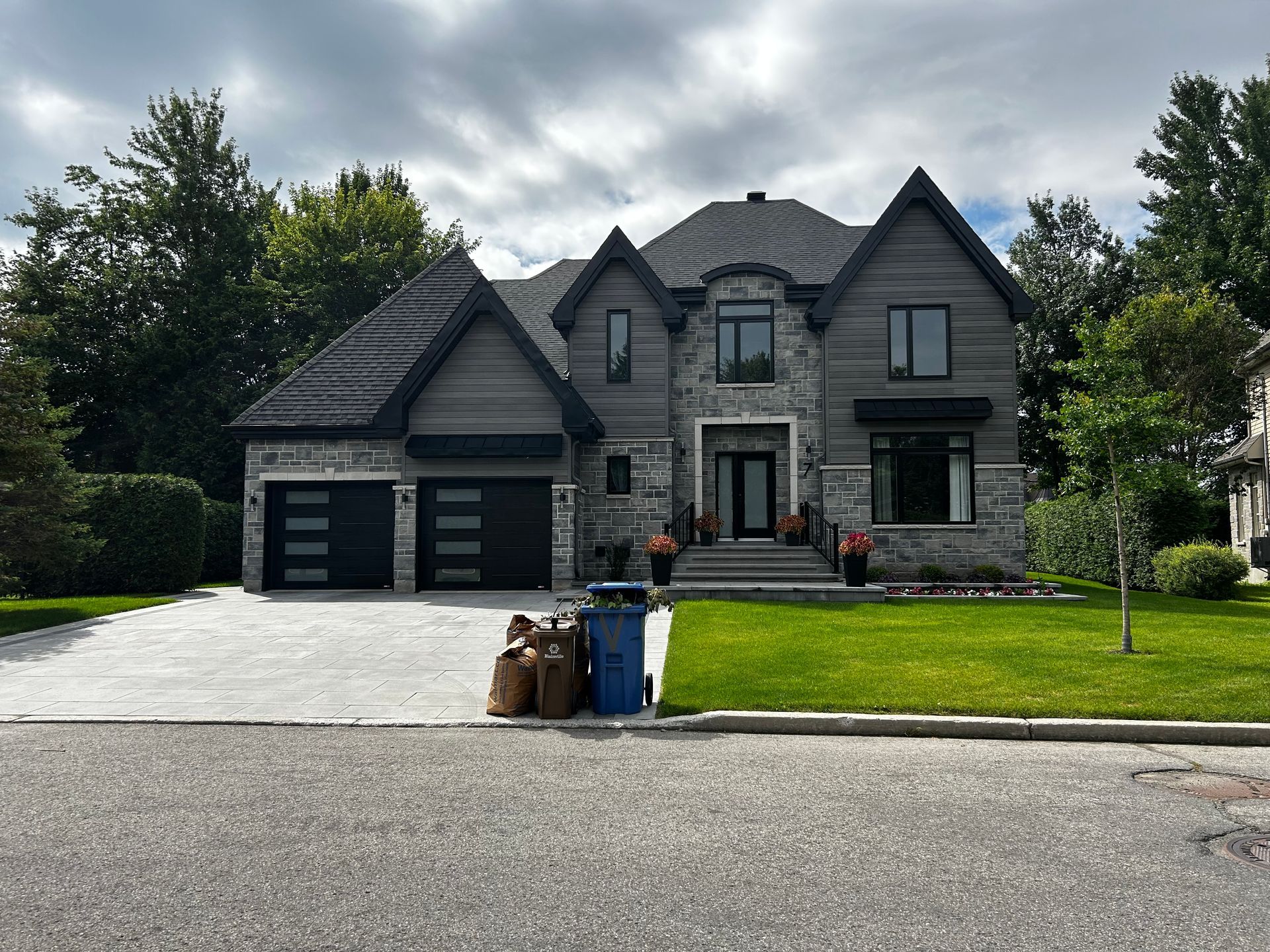 Maison en pierre grise avec portes de garage noires, pelouse verte et ciel nuageux.