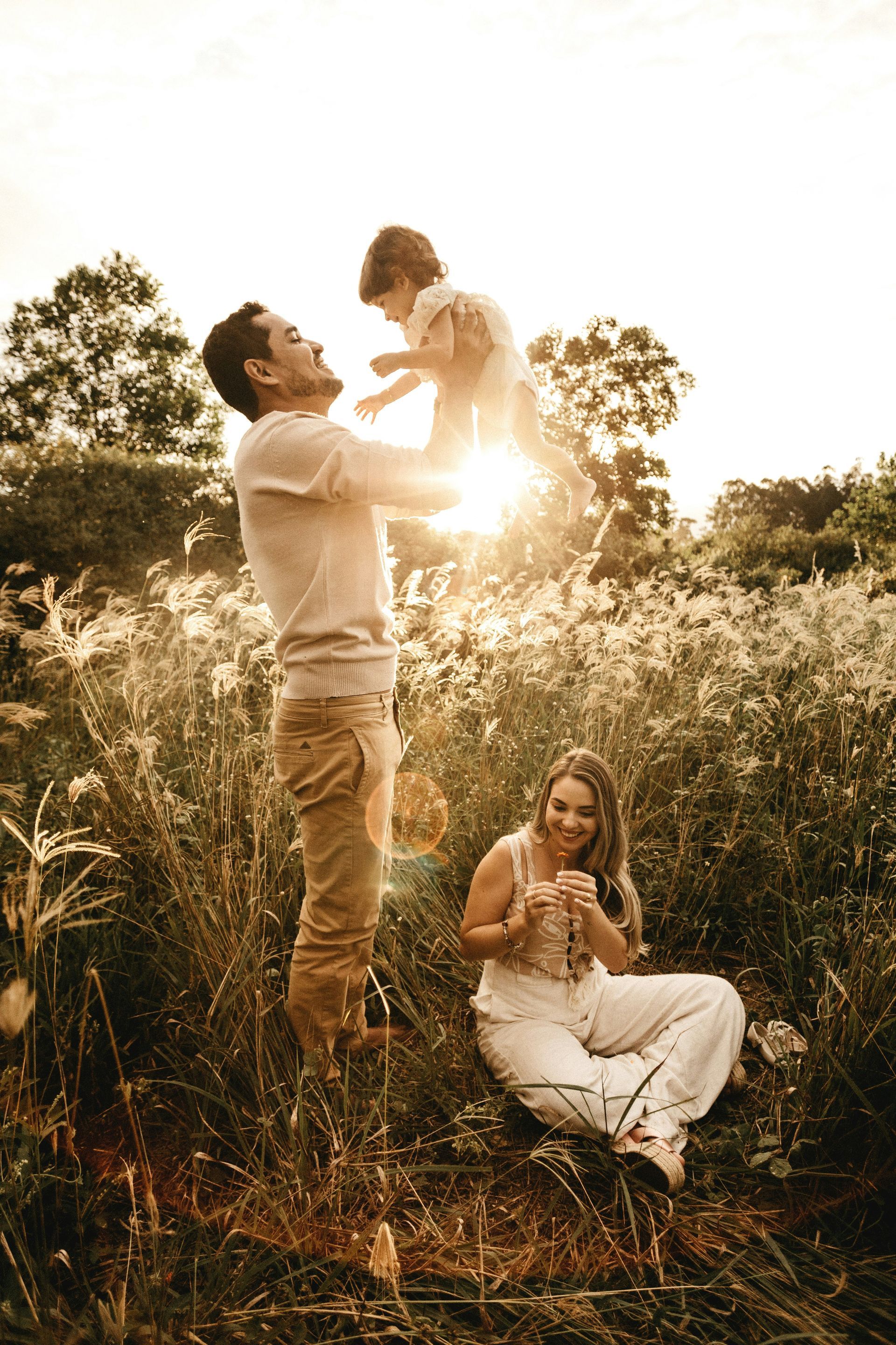 A man is holding a child in the air while a woman sits in the grass.