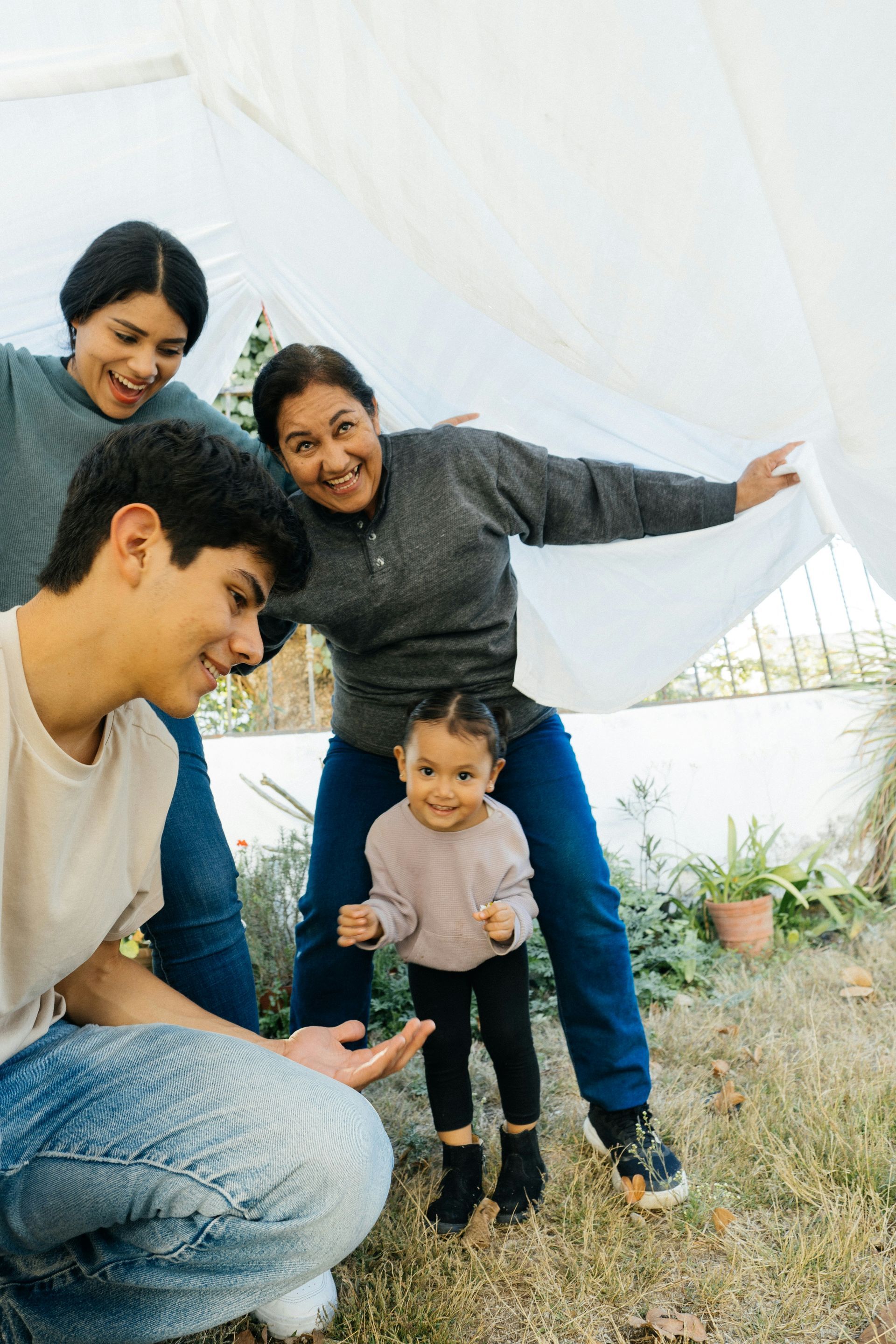 A family is playing under a white sheet in the grass.