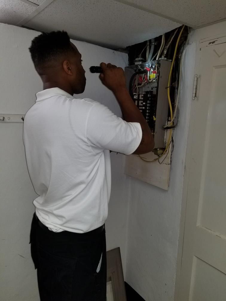 A man in a white shirt examines an electrical panel with a flashlight in a small room.