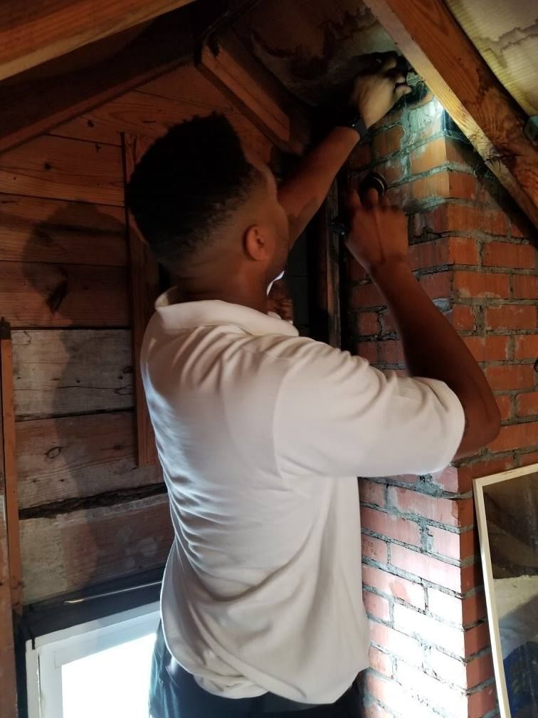 Man in white shirt inspecting in an attic, holding a flashlight. Brick wall and wooden beams visible.