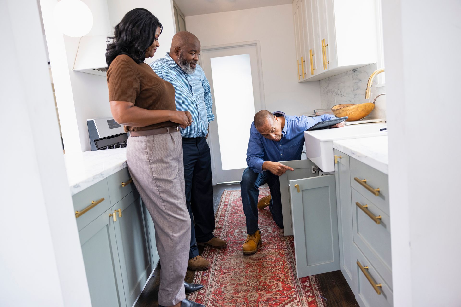 Couple watching a plumber kneeling under a kitchen sink.