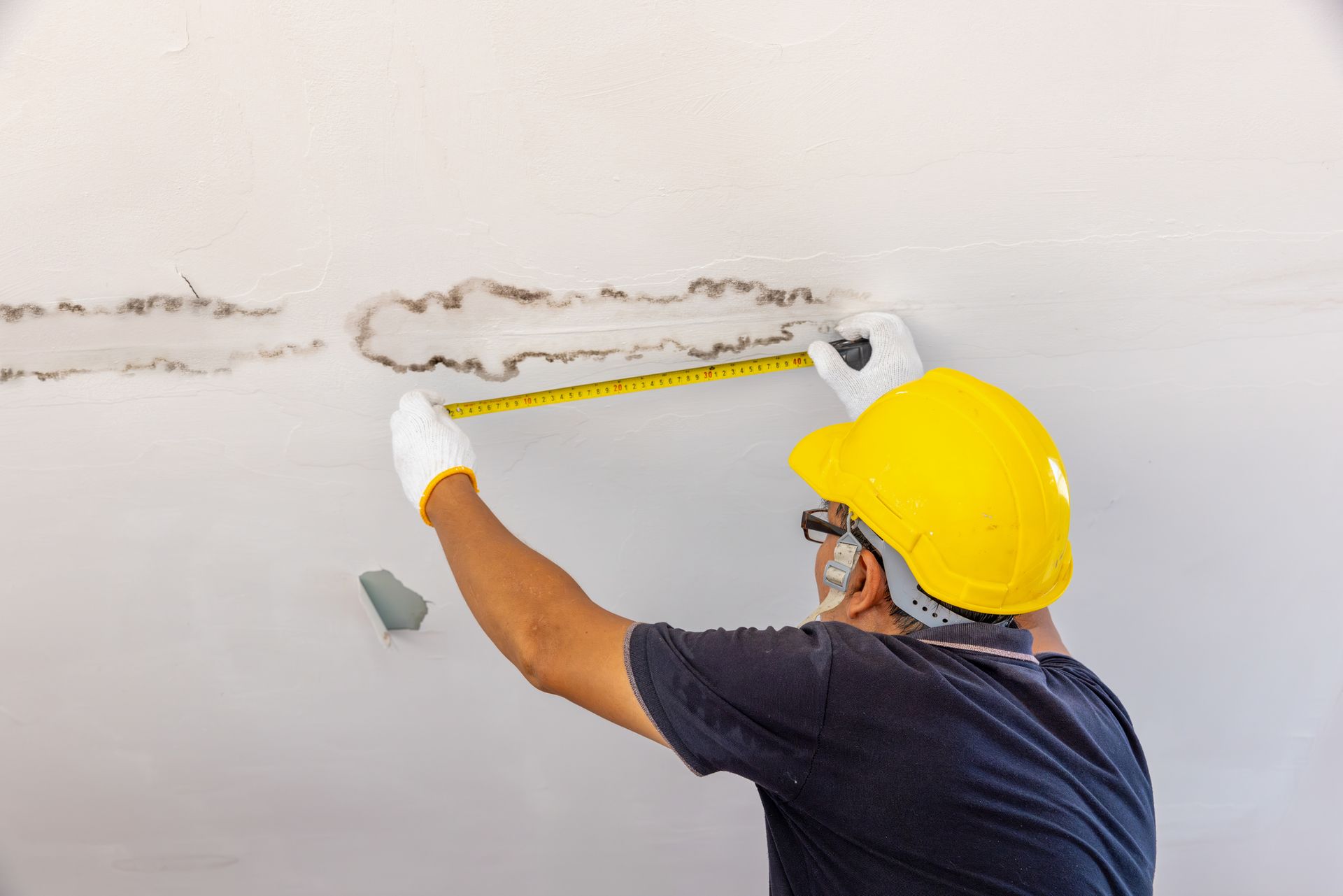 Person in a yellow hard hat measures water damage on a white ceiling, wearing gloves.