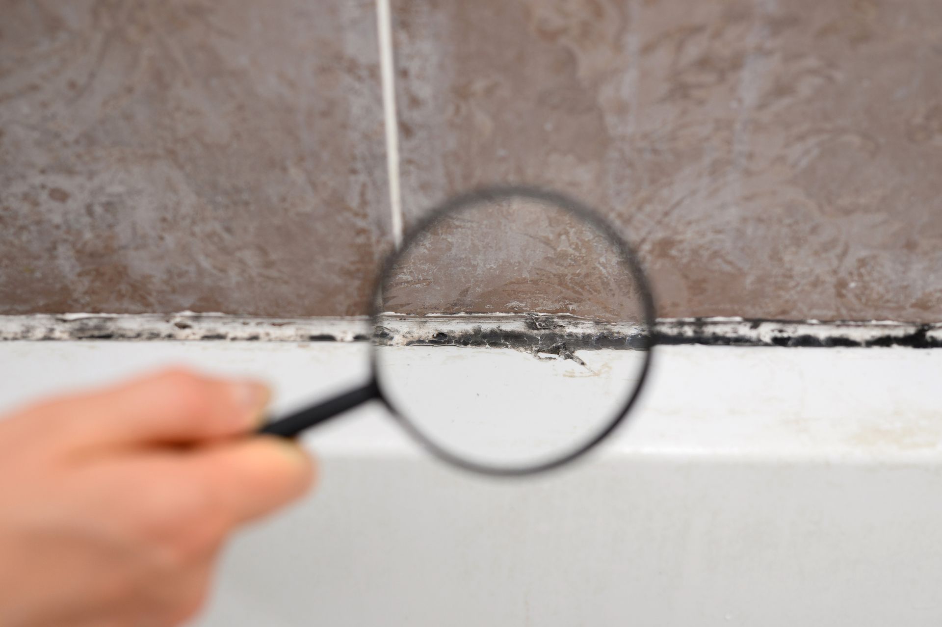 Hand holding a magnifying glass, examining black mold in a bathroom.
