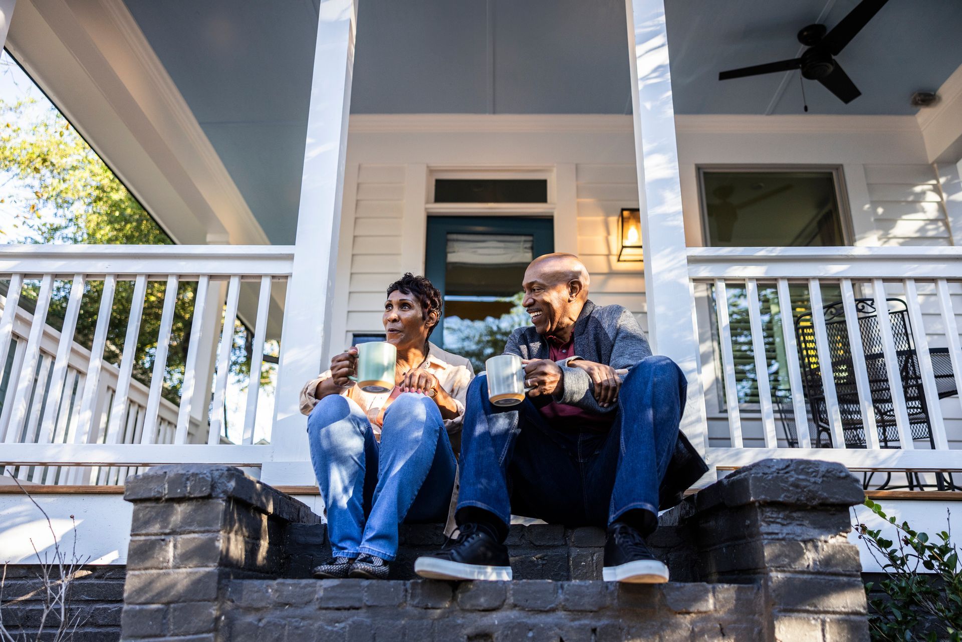 Two people sit on porch steps, holding mugs.