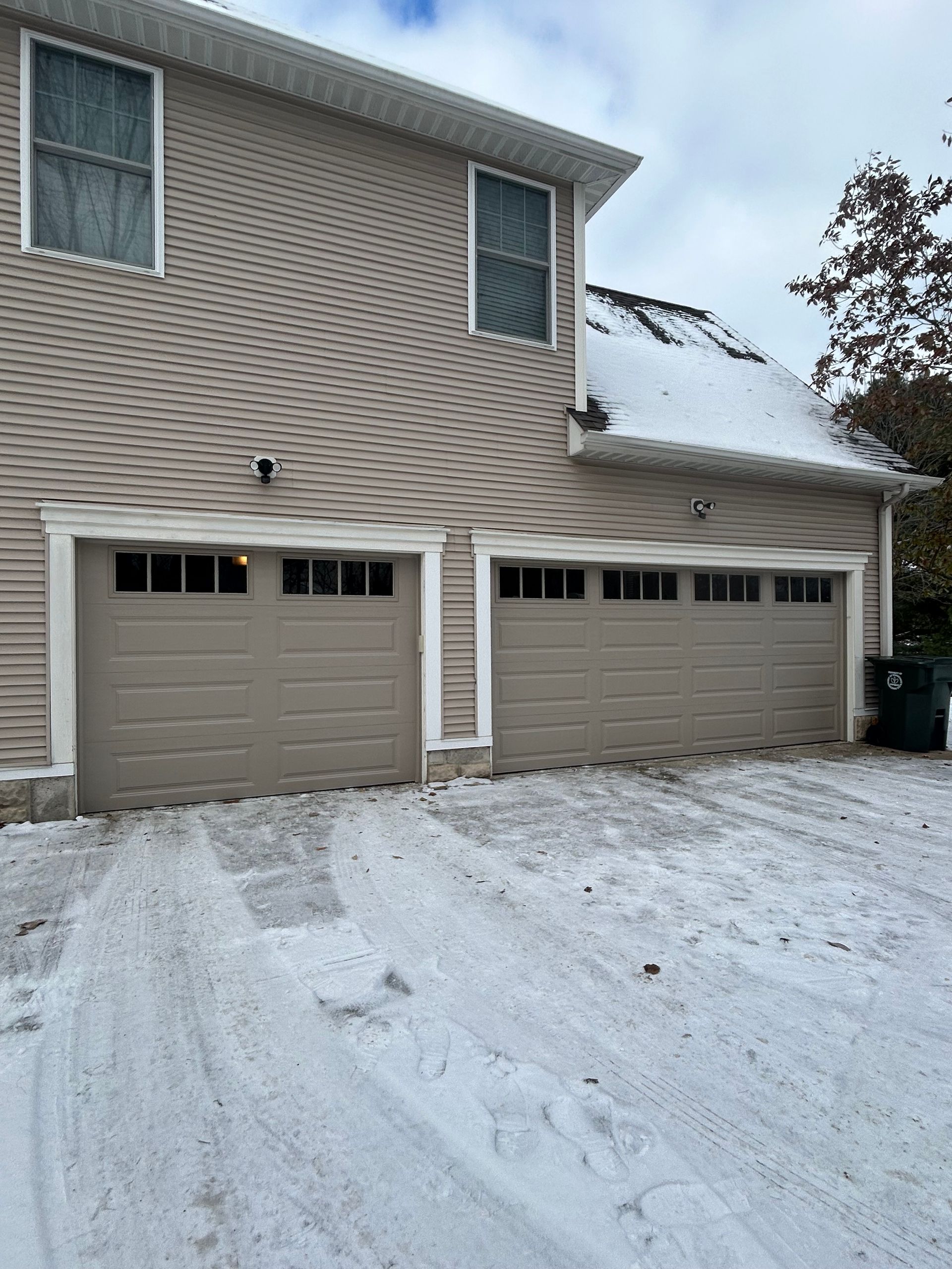 Two tan garage doors on a house with a snow-covered driveway and roof.