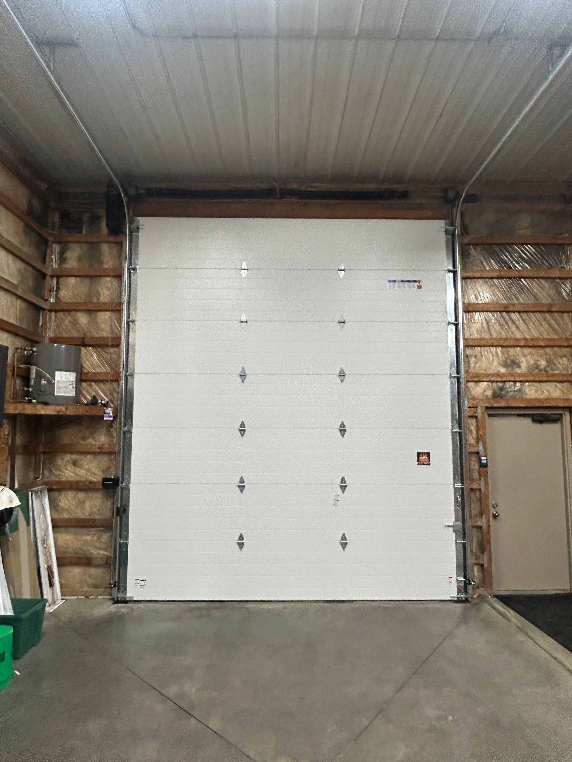 White garage door inside a building, flanked by wood and insulation. Concrete floor.
