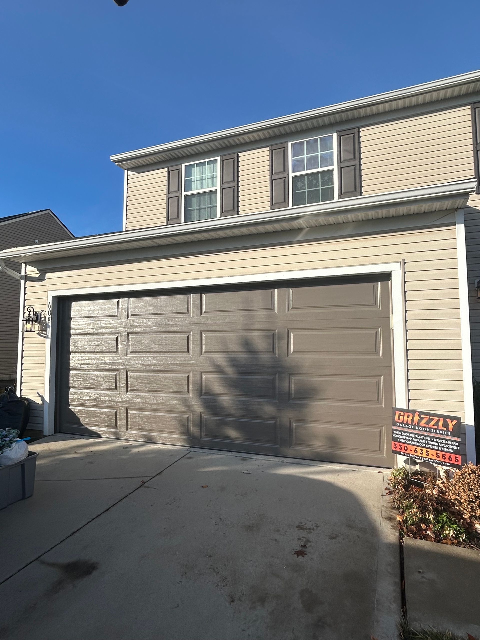 Garage door with peeling paint on a two-story house with dark shutters and blue sky.