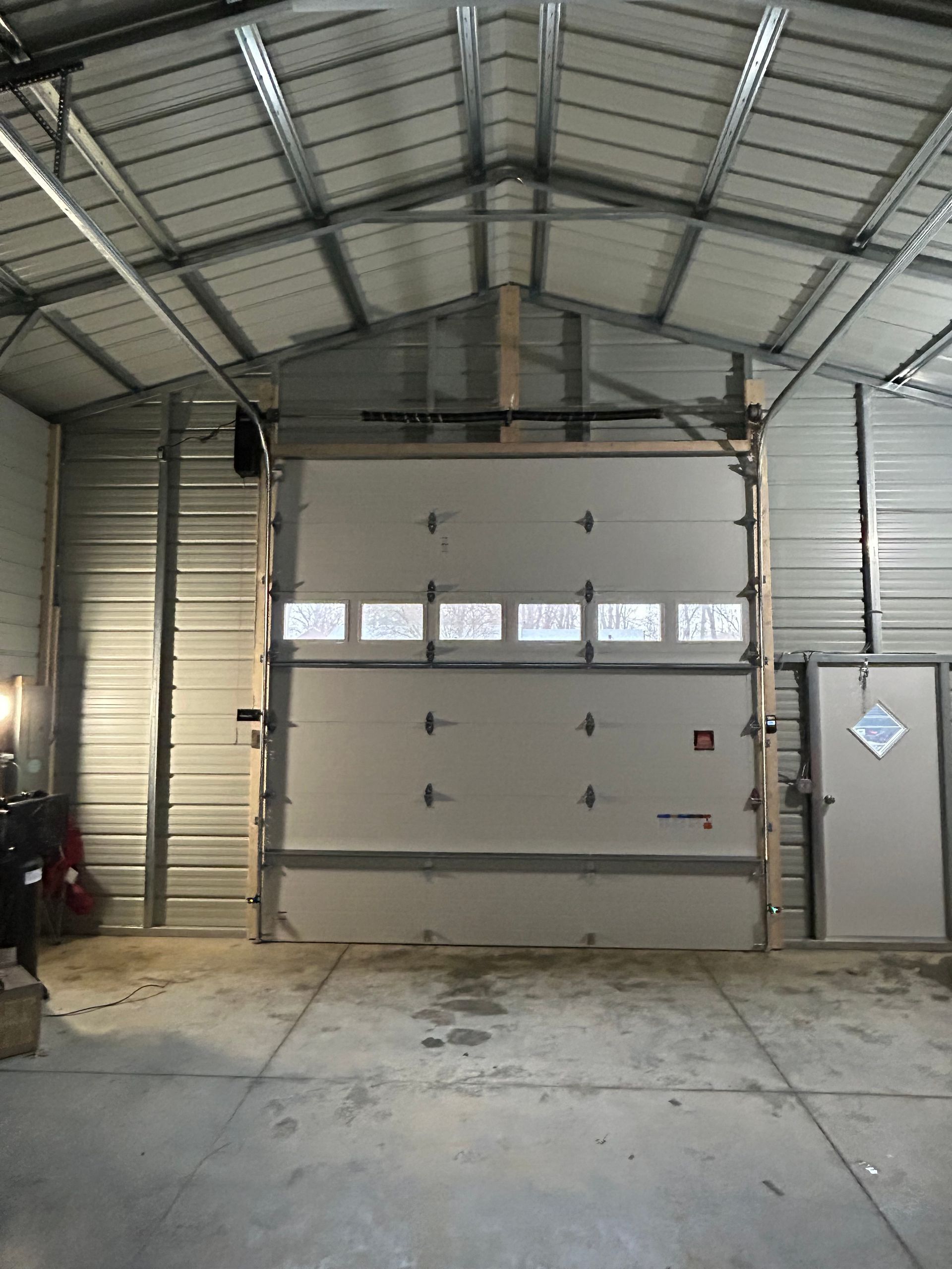 Interior of a metal building with a large garage door, a side door, and corrugated metal walls and ceiling.