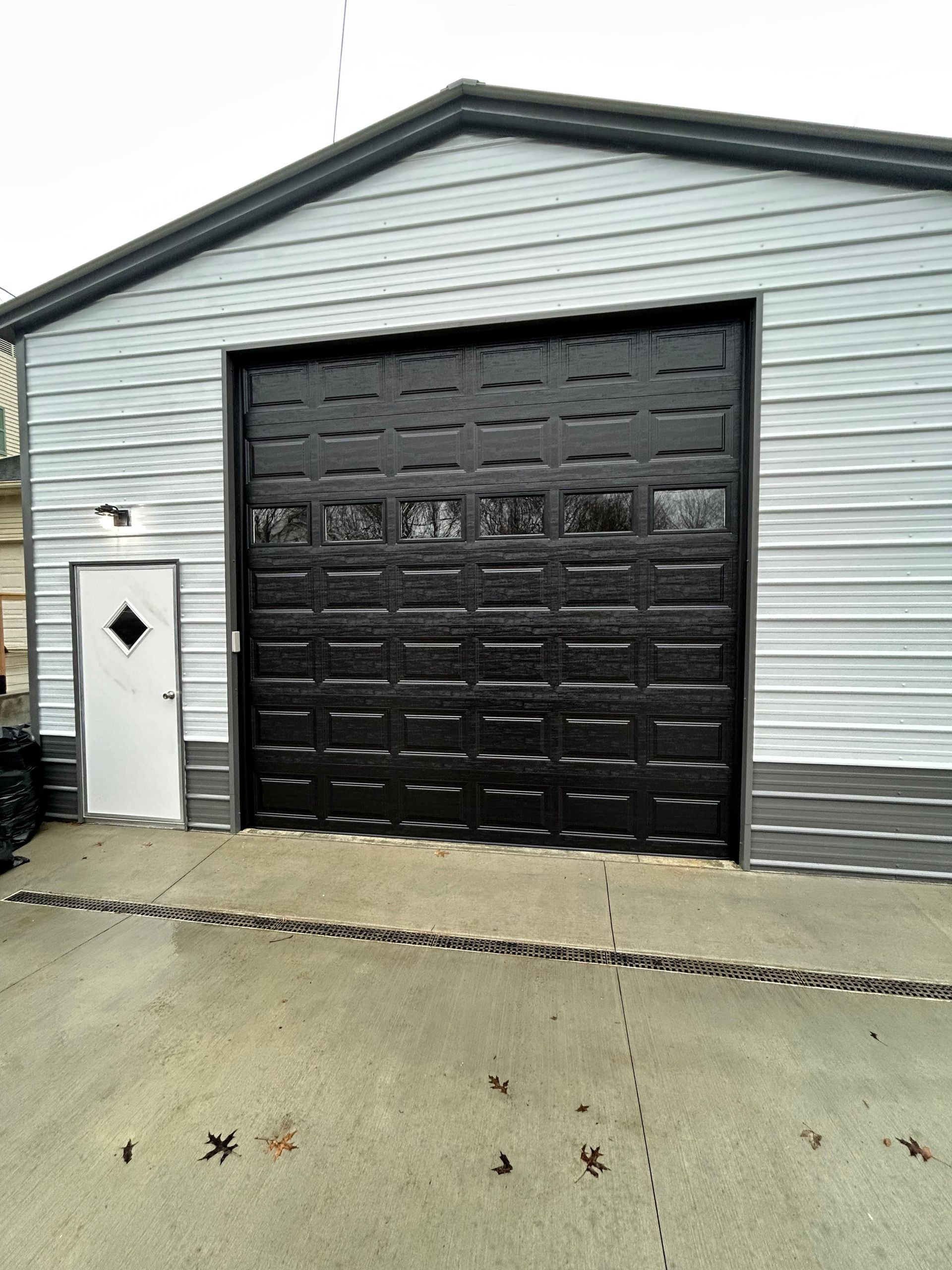 Metal garage with black overhead door and white side door. Gray siding with black trim on a concrete pad.