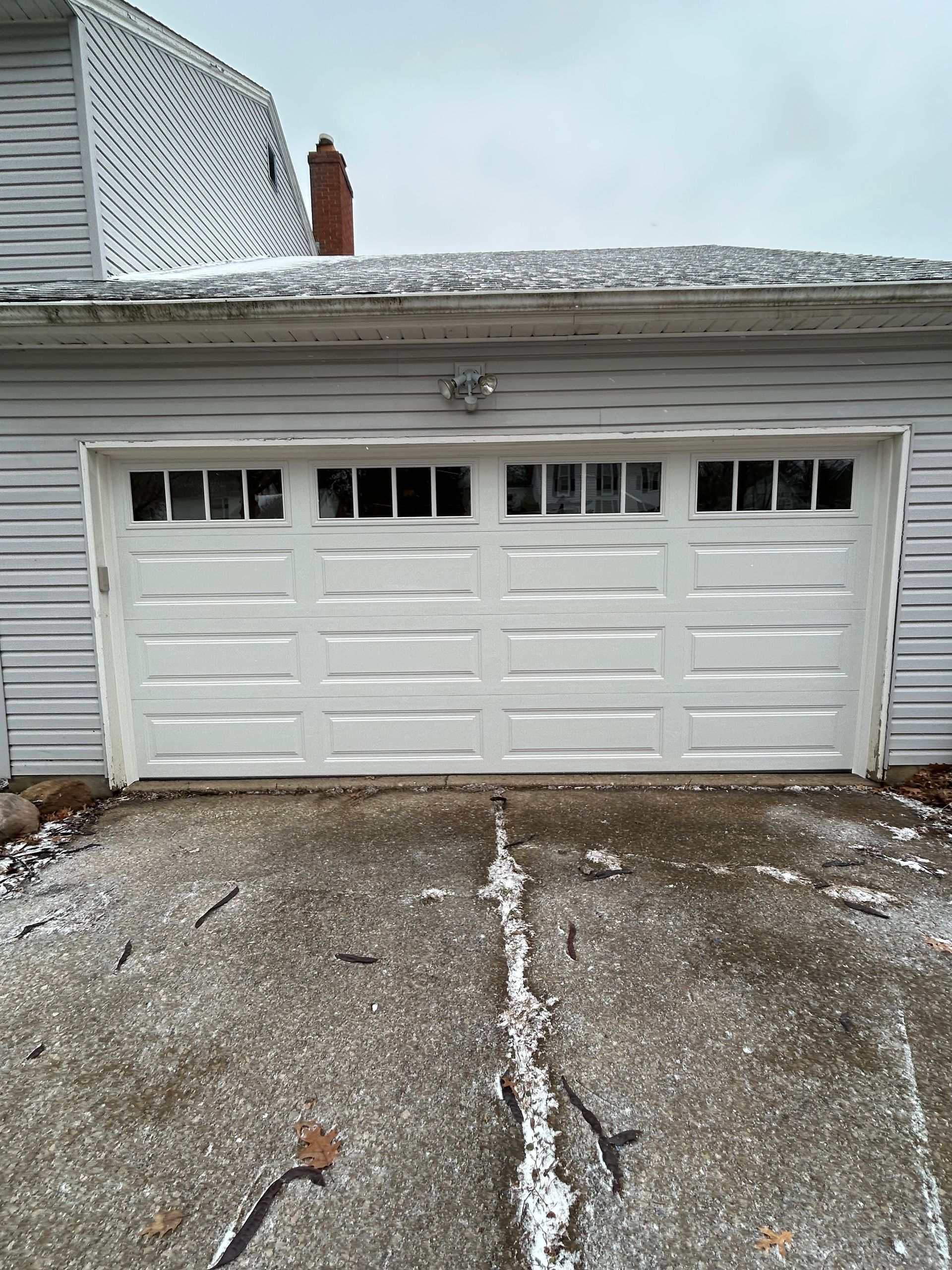 White garage door on a weathered, white house. Concrete driveway in front. Overcast day.
