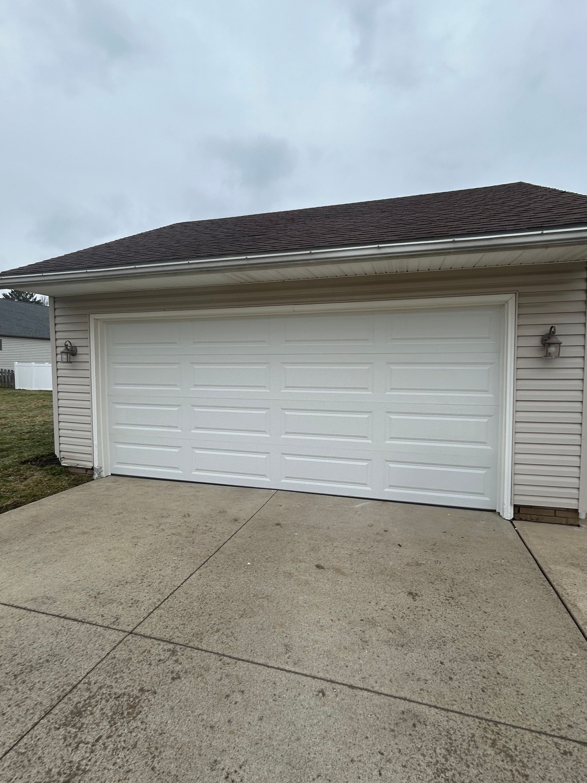 White garage door on a concrete driveway, attached to a house with light siding and a brown roof.