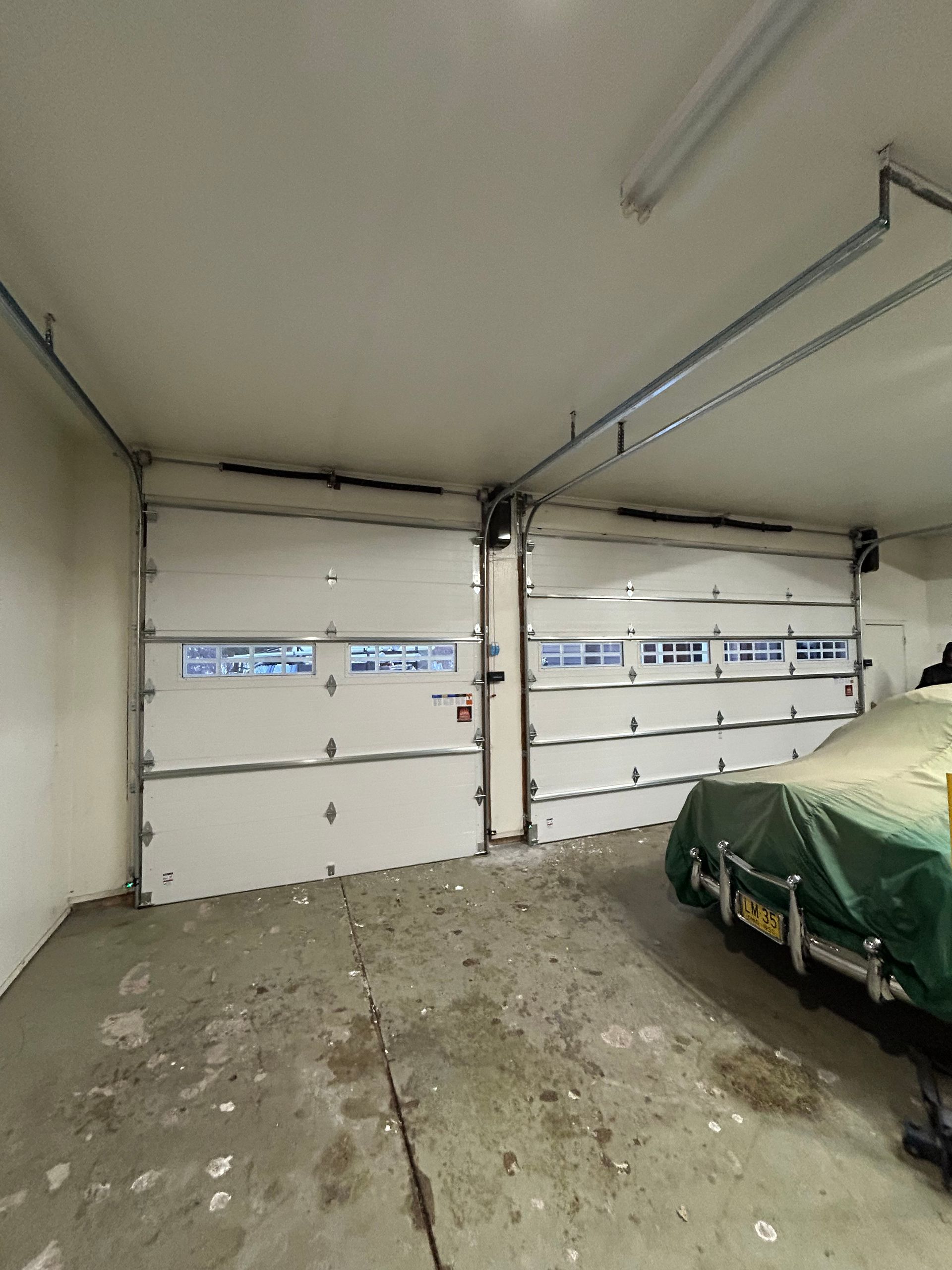 Two white garage doors with windows in a garage, vehicle covered on right.