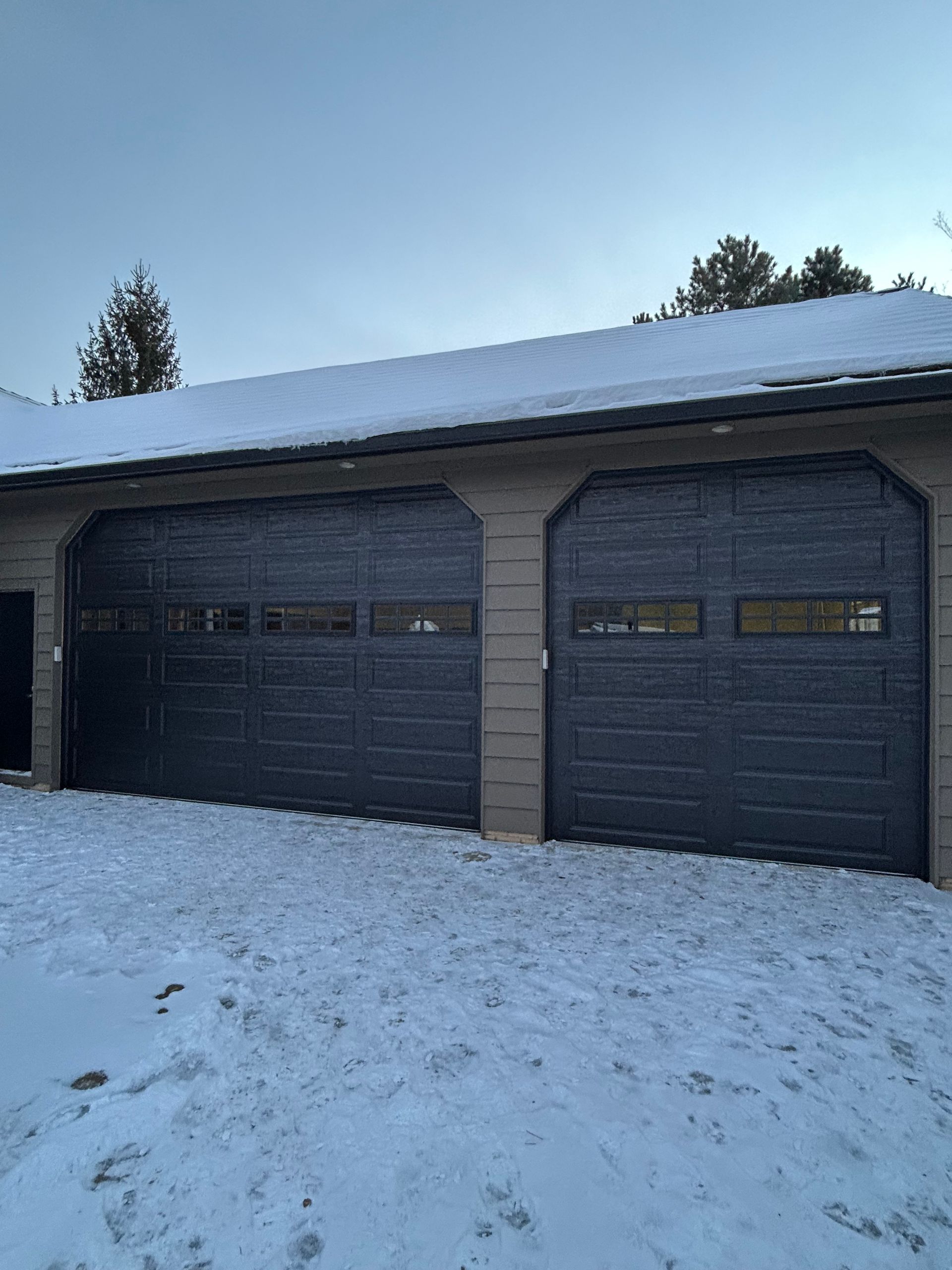 Two dark gray garage doors with small windows, covered in snow, under a snow-covered roof.
