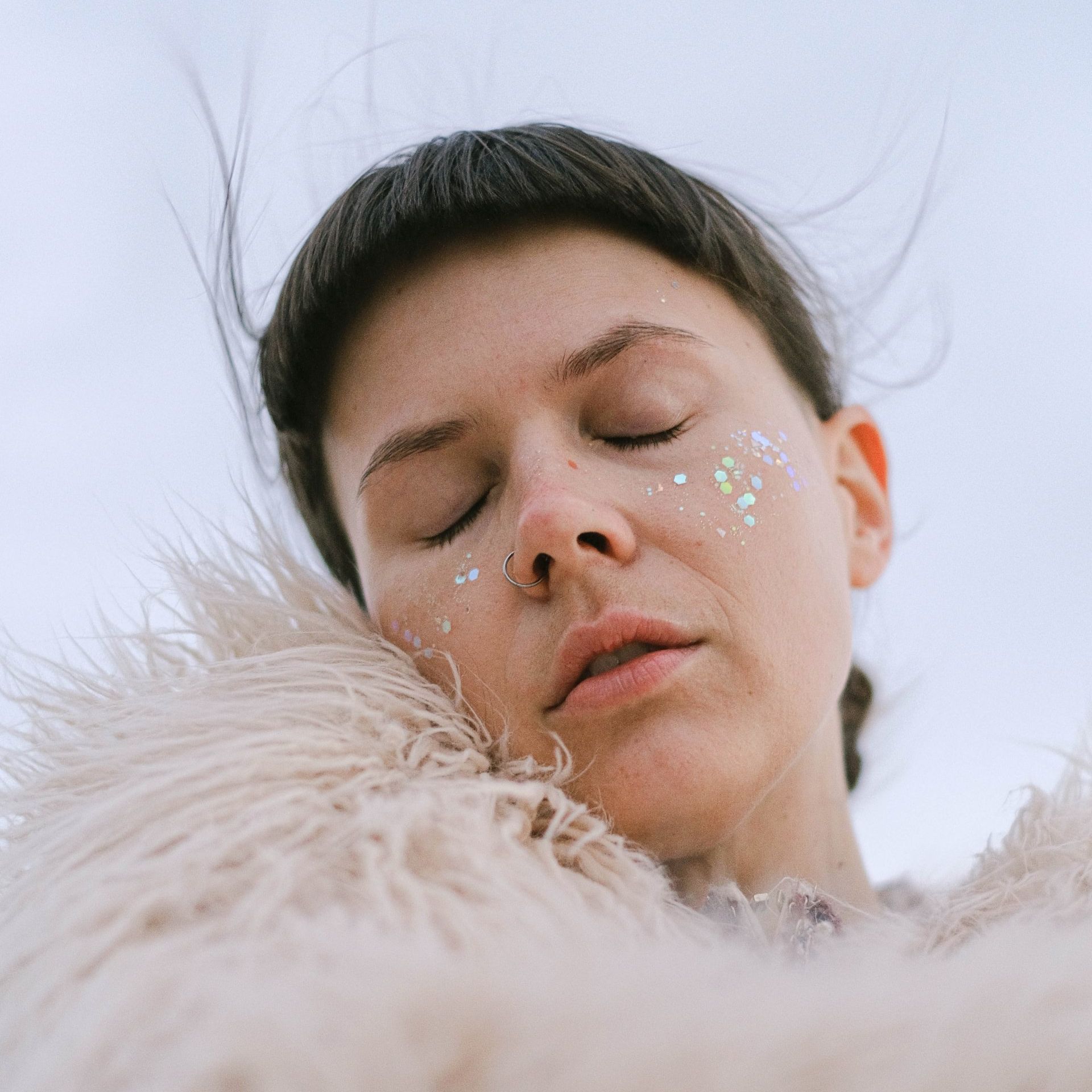 A woman with glitter on her face is laying on a furry blanket with her eyes closed.