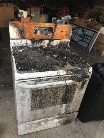 A dirty stove is sitting in a garage next to a trash can.