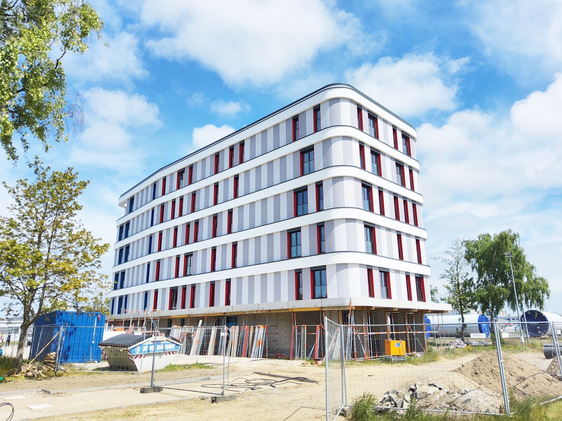 Ein großes modernes Gebäude mit weißen, grauen, roten und blauen Fassadenelementen und einem Bauzaun davor