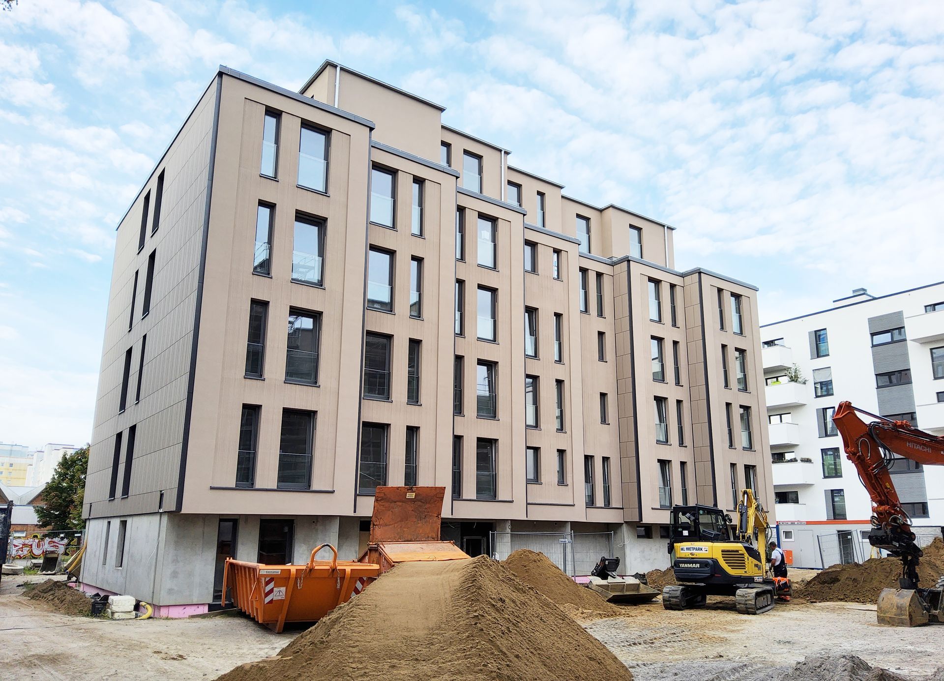 Ein großes Mehrfamilienhaus mit interessanter Architektur und Bagger, Container sowie Sandhaufen davor.