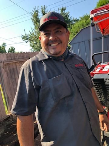 A smiling person in a gray uniform and cap stands outdoors near a wooden fence and red equipment on a sunny day.