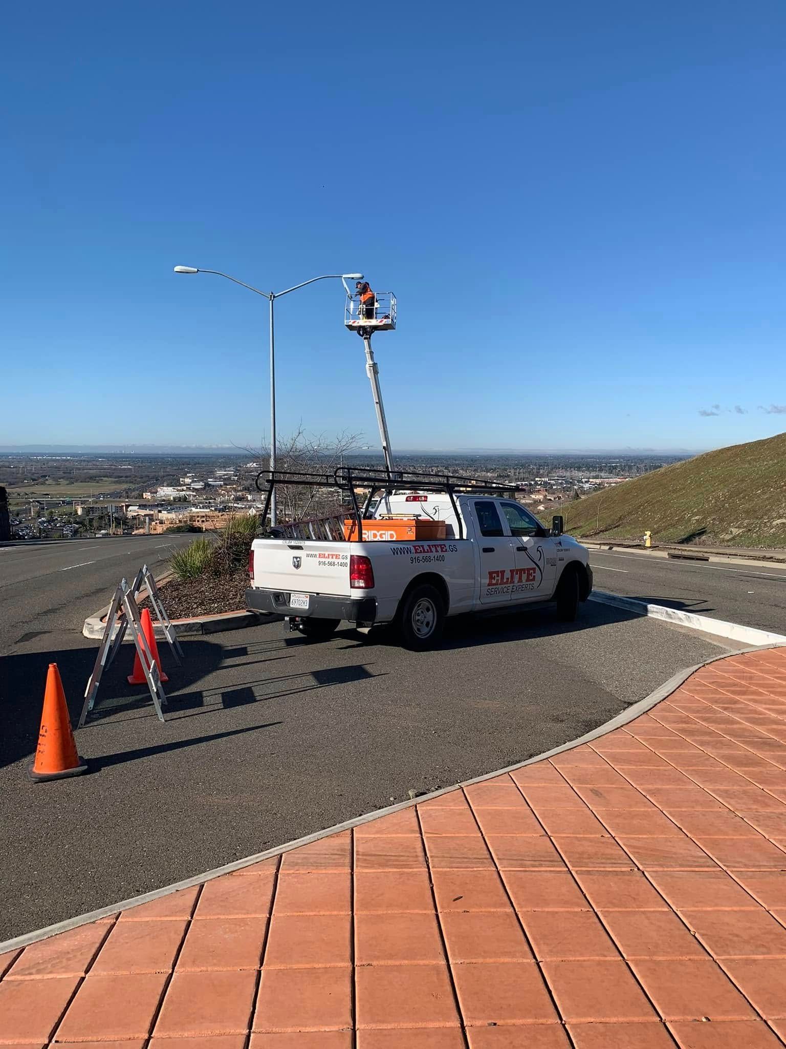 A worker in a bucket truck lifts to repair an outdoor light pole on a gravel hill overlooking a town.