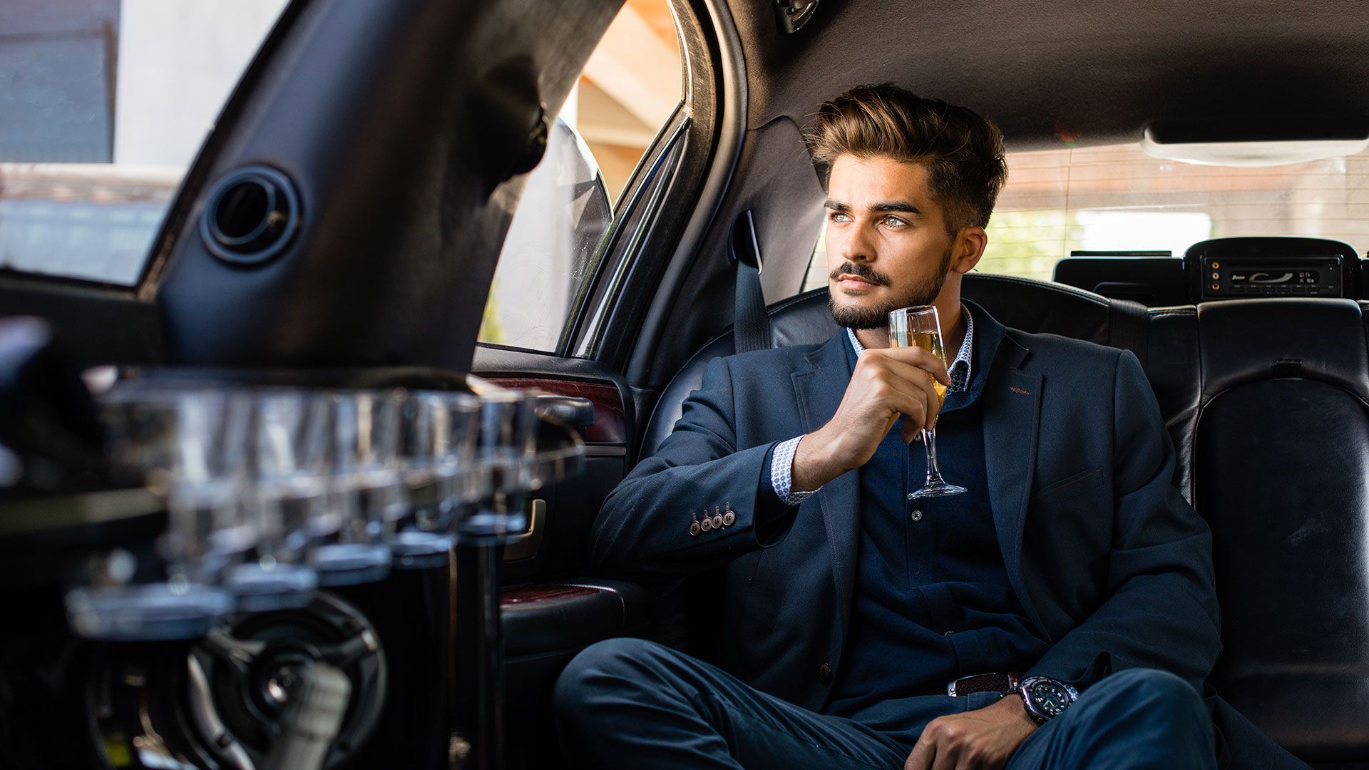 A man is sitting in the back seat of a limousine drinking a glass of wine.