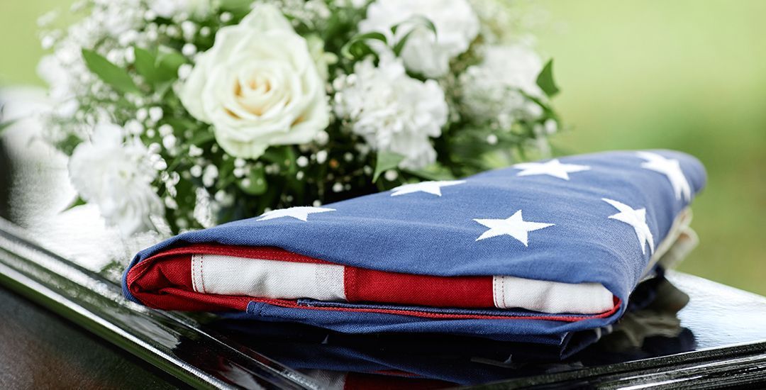 An American flag is lying on a casket with a bouquet of white roses.