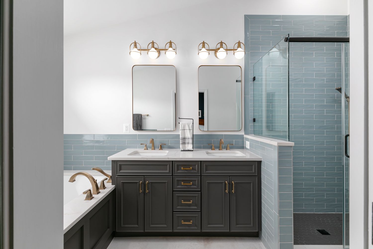 Modern bathroom with gray vanity, two mirrors, and a blue-tiled shower, lit by brass sconces.