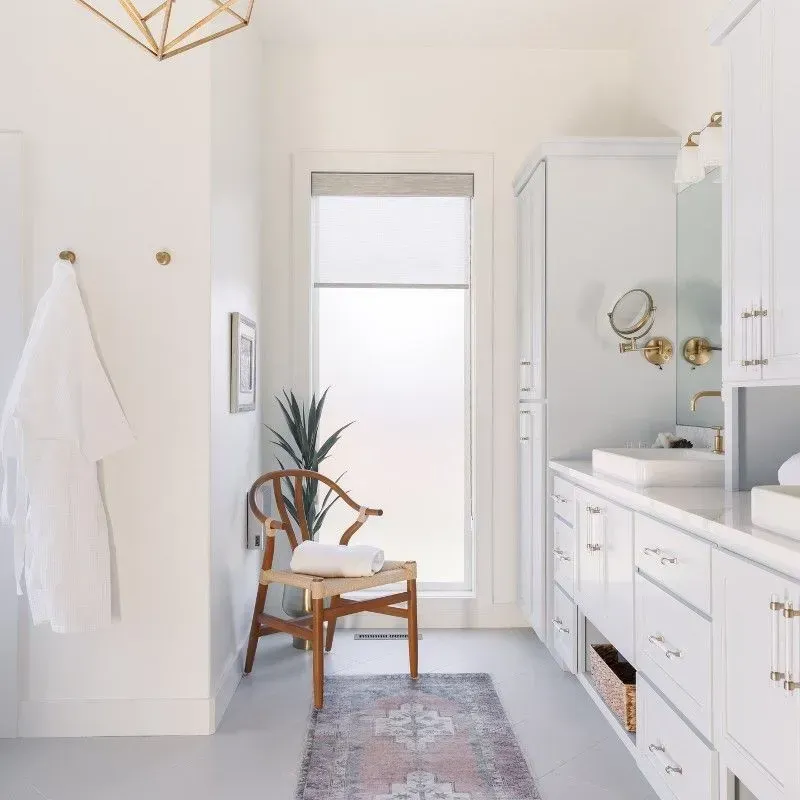 White bathroom with a chair, window, cabinets, and a decorative rug.