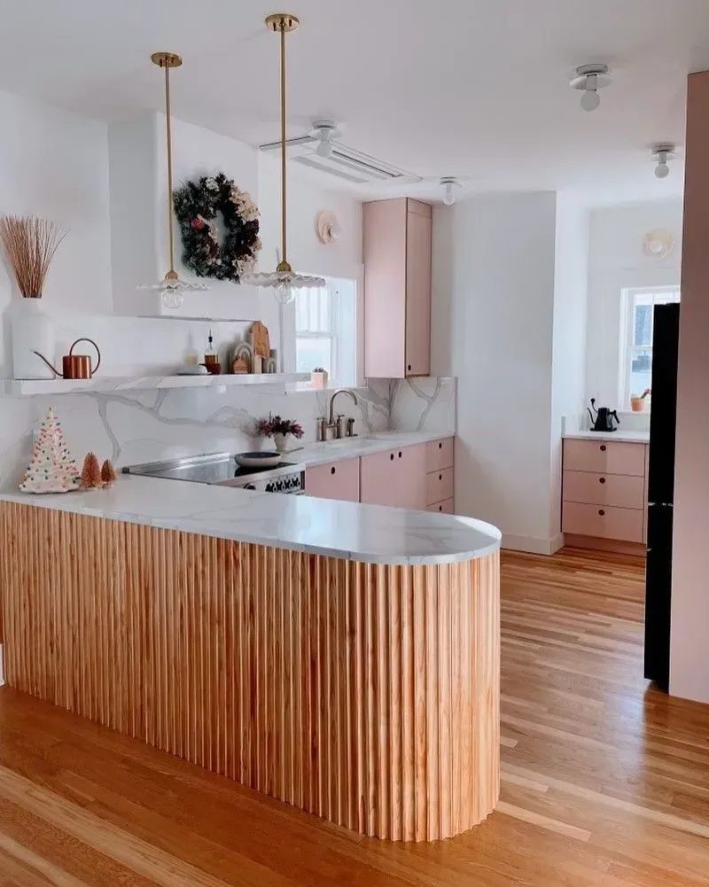 Modern kitchen with wood island, pink cabinets, marble countertops, and light fixtures.