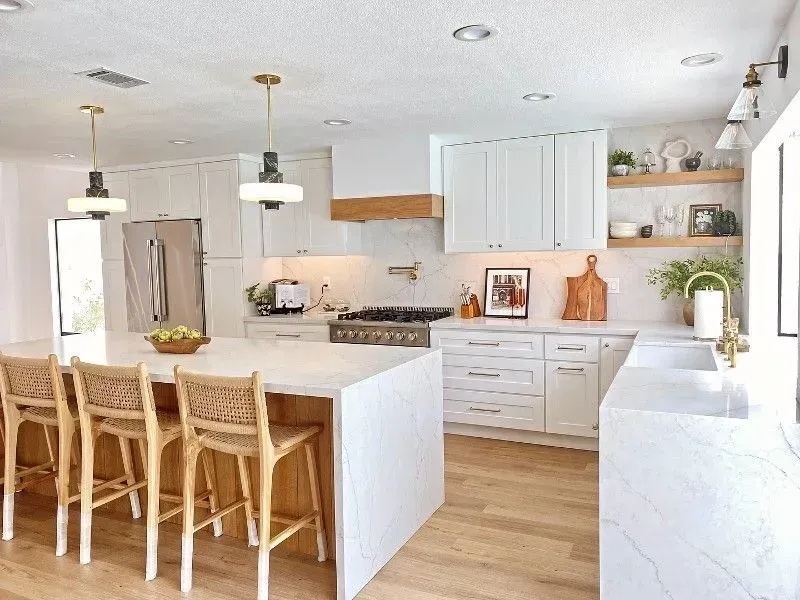 Modern white kitchen with island, wooden stools, and brass accents; stainless steel appliances.