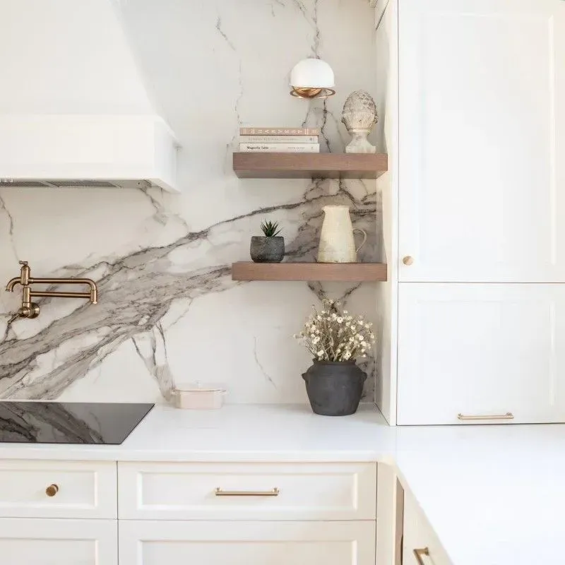 Modern kitchen with white cabinets, marble backsplash, and wooden shelves displaying decor.