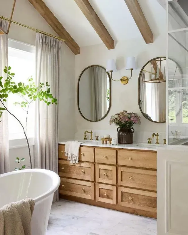 Bathroom with wooden vanity, oval mirrors, and white tub. Beige walls, light drapes, and beams.