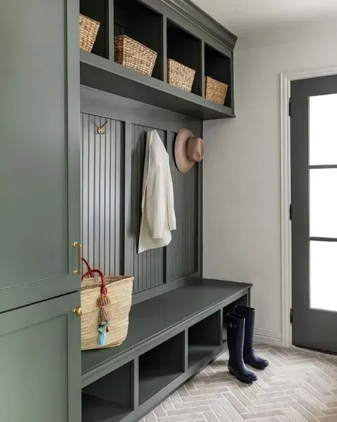Green built-in entryway storage with bench, hooks, and baskets. Boots, hat, and coat hanging. Herringbone floor.