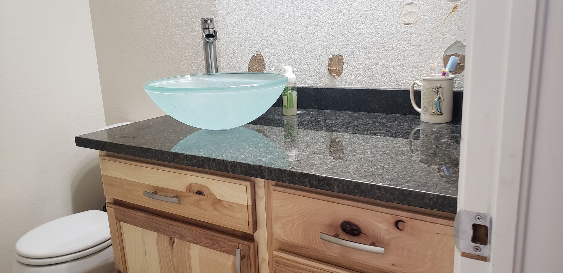Bathroom vanity with glass bowl sink and dark countertop, wooden cabinets, and a decorative backsplash.