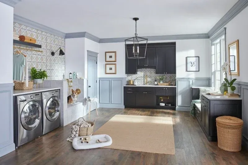Laundry room with washing machines, cabinets, dog bed, and a beige rug. Light blue and white walls.