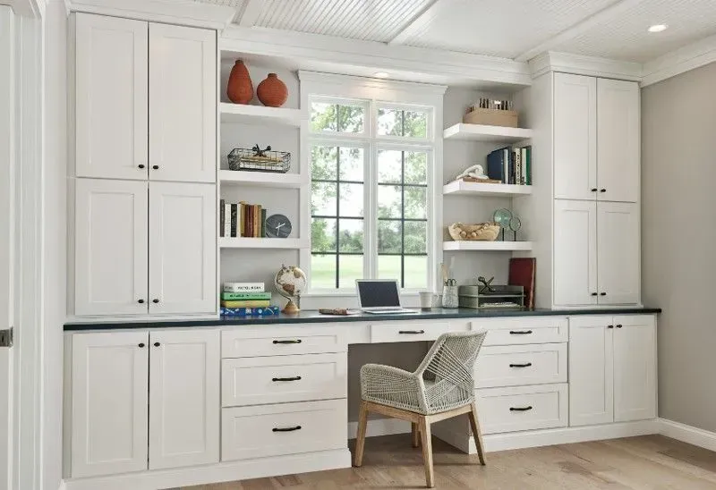 Built-in white cabinets with desk and window. Decorative shelves with books and decor. Neutral walls, wood floor.