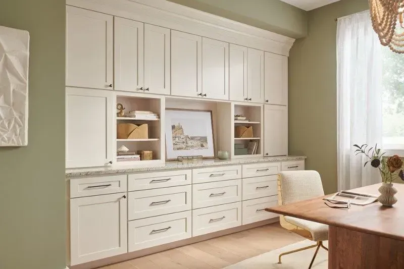 White cabinets with drawers and shelving in a home office with a desk and window.