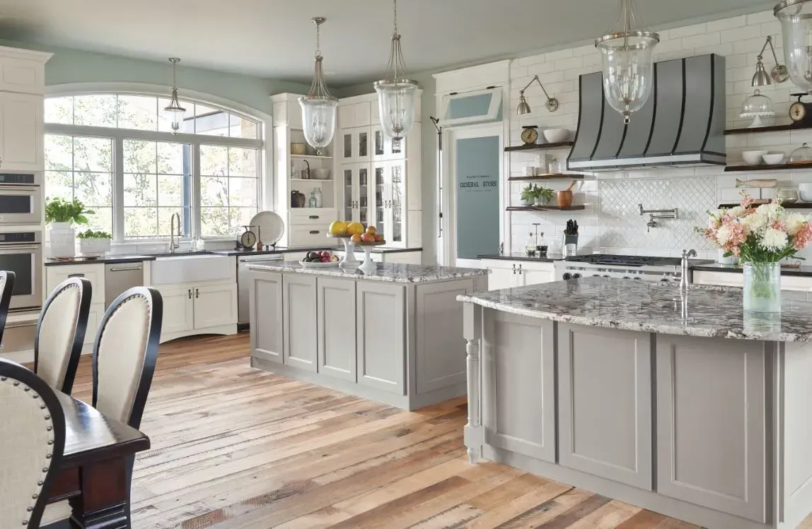 Spacious kitchen with gray cabinets, marble countertops, and light wood flooring. Large arched window and pendant lights.