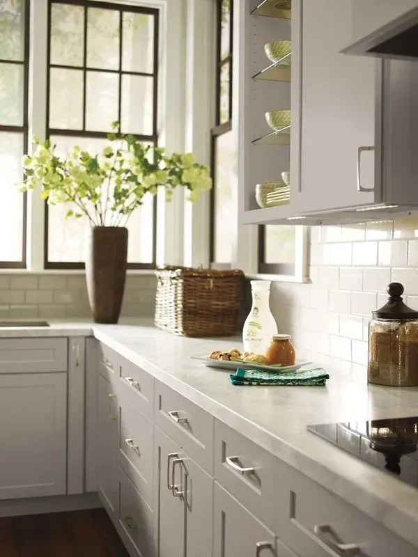 White kitchen countertop with cabinets, backsplash, and windows; a vase of flowers, and various kitchen items.