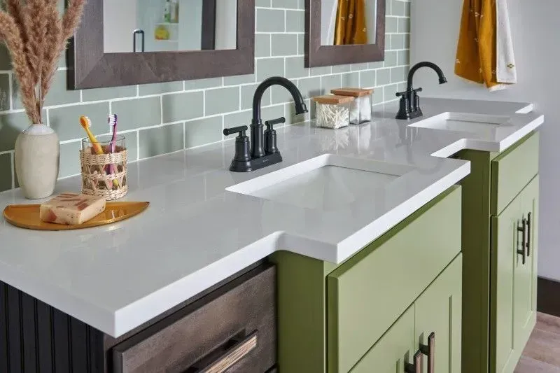 Bathroom with green cabinets, white countertop, black faucets, and sage green tile backsplash.