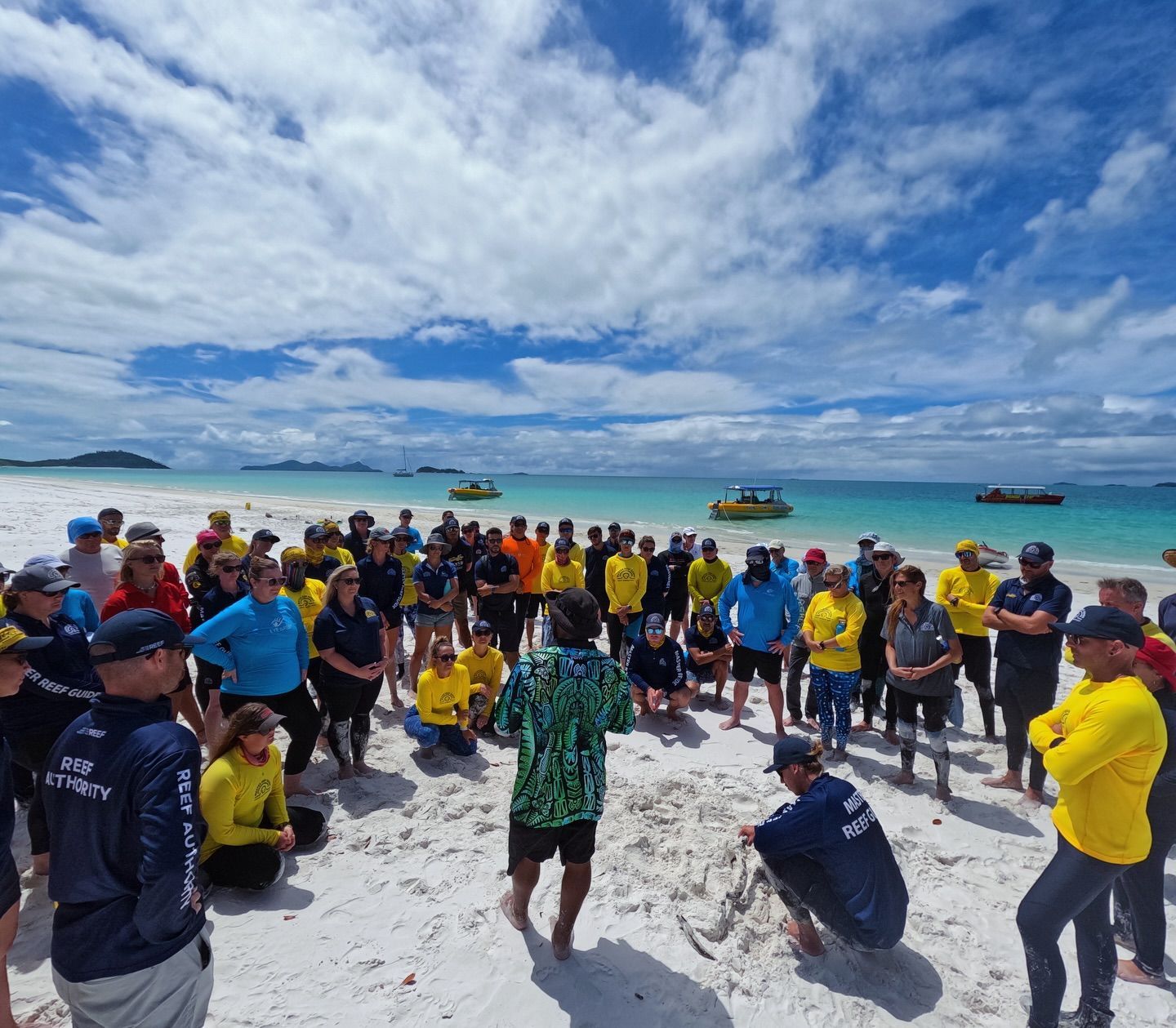 A group of people are gathered on a beach with boats in the background