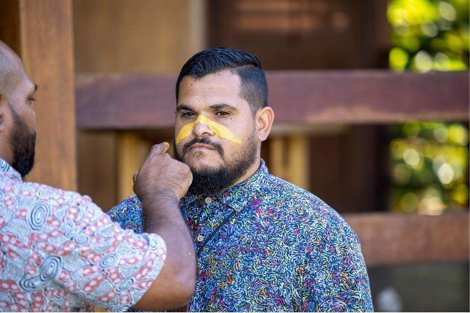 A man with a beard is getting his face painted by another man.