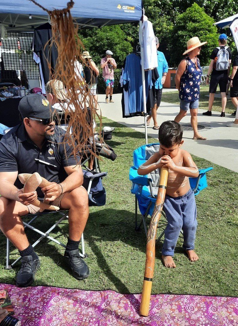 A little boy is playing a didgeridoo while a man sits in a chair.