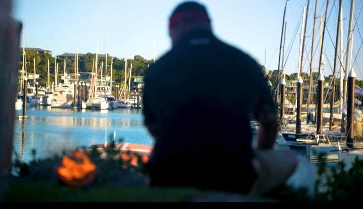 A man in a black jacket is standing in front of a harbor filled with boats.