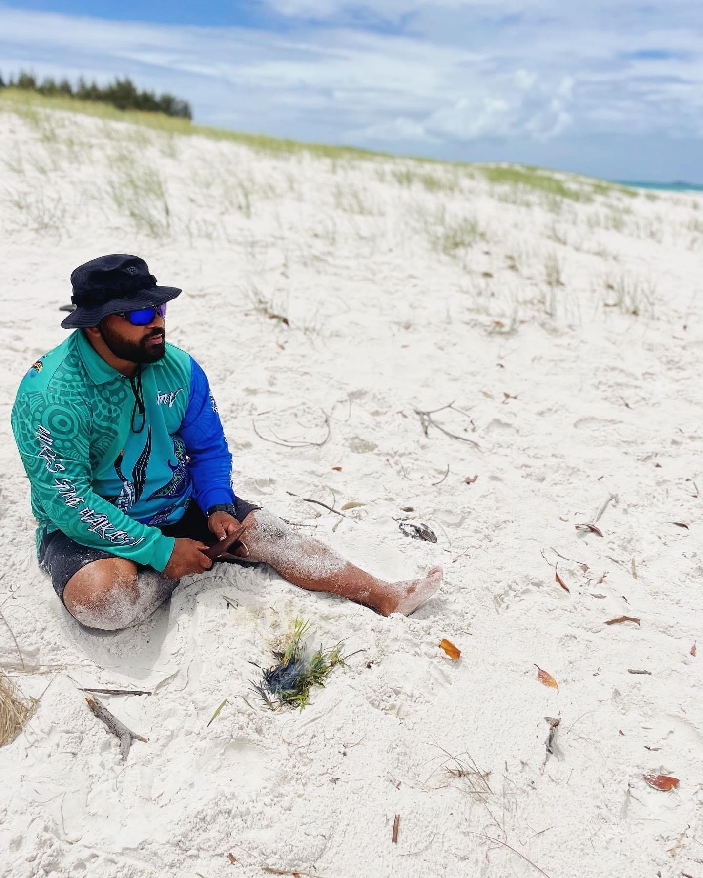 A man is sitting on a sandy beach wearing sunglasses and a hat.