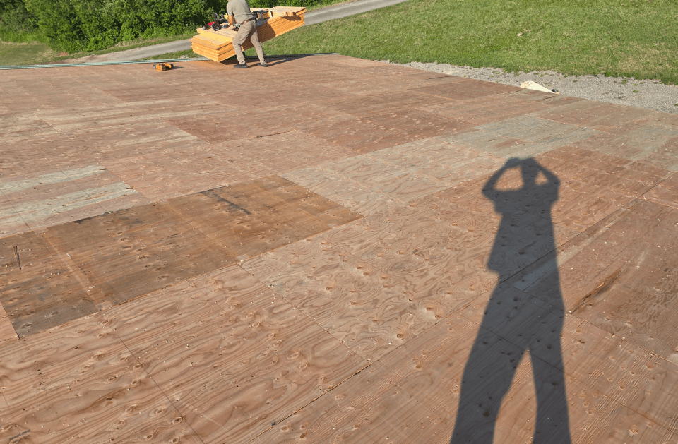 Person's shadow on reddish-brown wooden boards. Another person holds an orange tool on the wooden surface.