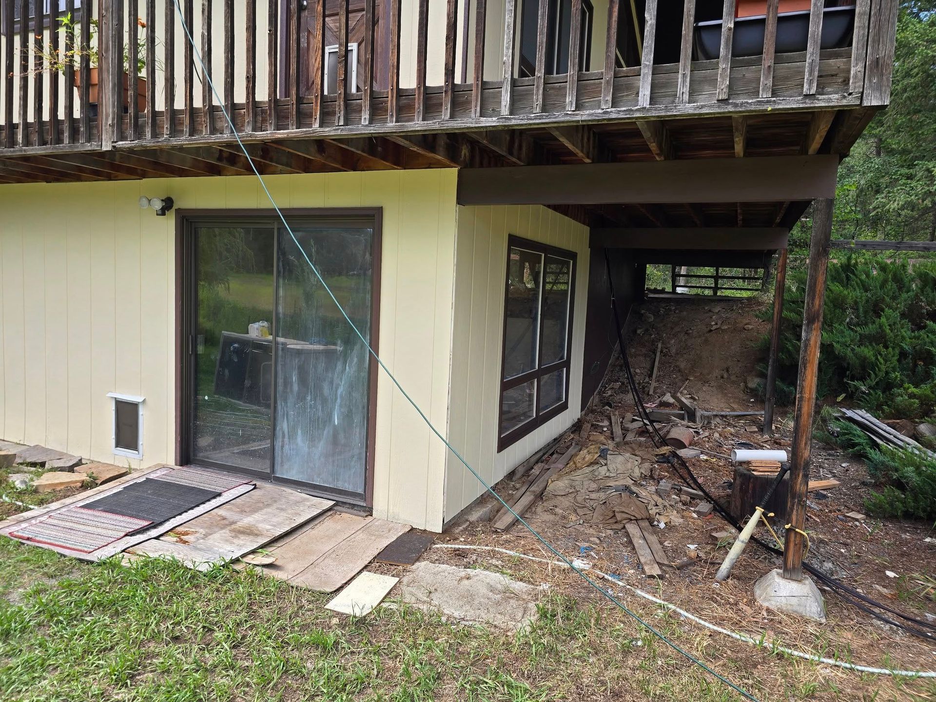 Beige building with sliding glass door under a wooden deck. Debris is present near the foundation.