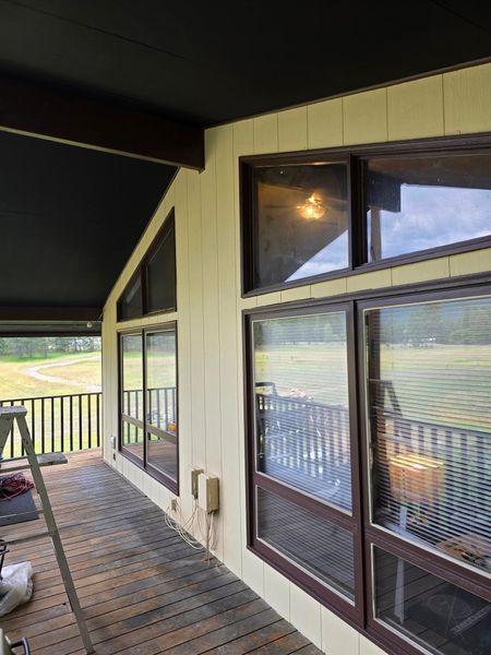 Deck with windows on a beige wall under a dark brown roof. Brown window frames, wooden deck, and railing.